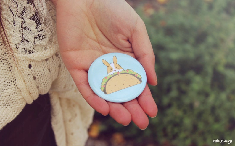 May include: A light blue button with a cartoon bunny rabbit wearing a taco. The taco is filled with lettuce, cheese, and meat.
