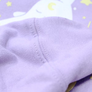 May include: Close-up of the purple fabric of a sweatshirt with a white narwhal design. The fabric is soft and textured.