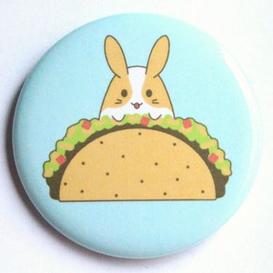 May include: A light blue button with a cartoon image of a brown and white bunny rabbit peeking out of a yellow taco with green lettuce and red tomato filling.
