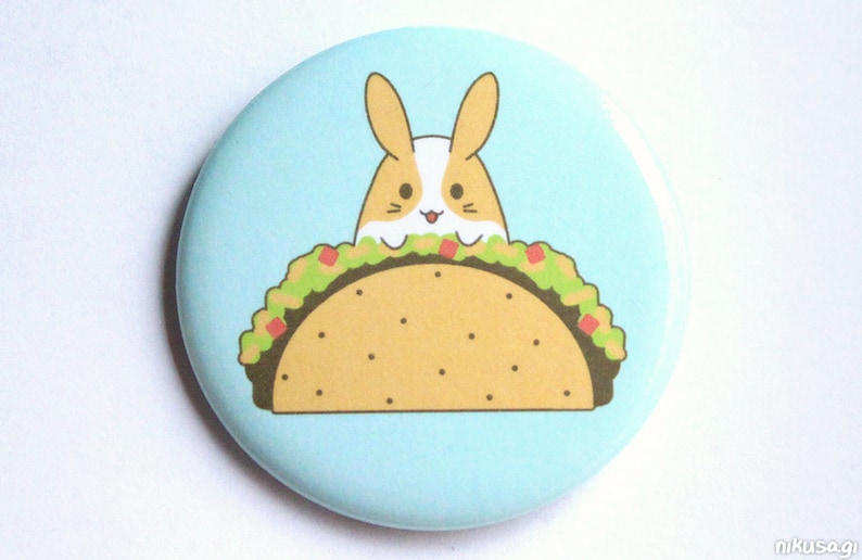 May include: A light blue button with a cartoon image of a brown and white bunny rabbit peeking out of a yellow taco with green lettuce and red tomato filling.