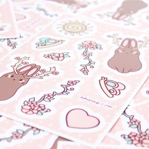 Może przedstawiać: Zestaw r&oacute;żowych i brązowych naklejek przedstawiających uroczego kr&oacute;liczka z rogami, kwiatami i sercem. Naklejki znajdują się na r&oacute;żowym tle z białymi kropkami. Na jednej z naklejek znajduje się napis "nakusagi.com".
