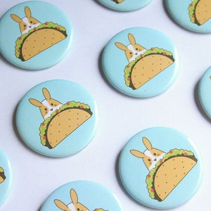 May include: A set of 10 round blue and white buttons with a cartoon bunny rabbit inside a taco. The bunny is white with pink ears and a pink nose. The taco is yellow with green lettuce and red tomato filling.