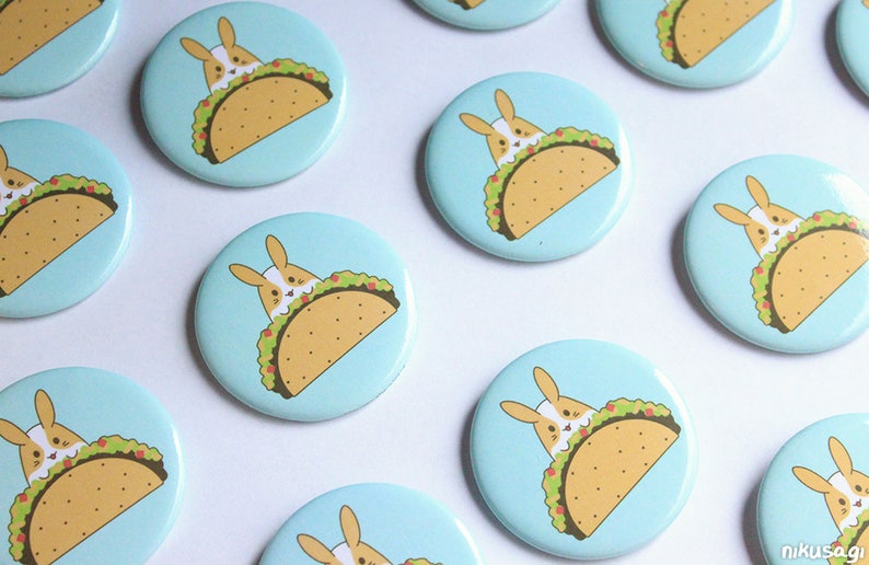 May include: A set of 10 round blue and white buttons with a cartoon bunny rabbit inside a taco. The bunny is white with pink ears and a pink nose. The taco is yellow with green lettuce and red tomato filling.