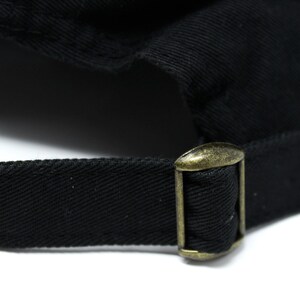 May include: Close-up of a black baseball cap with a brass-colored metal buckle and adjustable strap. The cap is made of a textured fabric, and the buckle is rectangular with a sliding mechanism.
