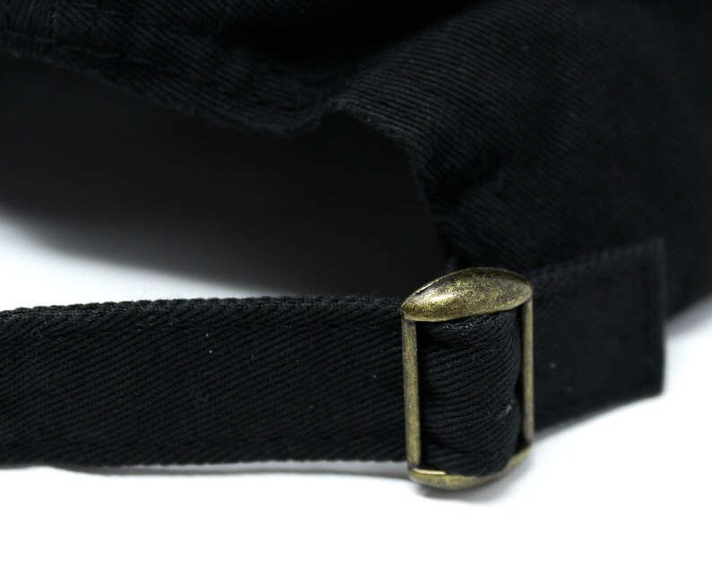 May include: Close-up of a black baseball cap with a brass-colored metal buckle and adjustable strap. The cap is made of a textured fabric, and the buckle is rectangular with a sliding mechanism.