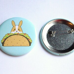 May include: A light blue button with a cartoon bunny rabbit peeking out of a taco. The back of the button is silver with a pin.
