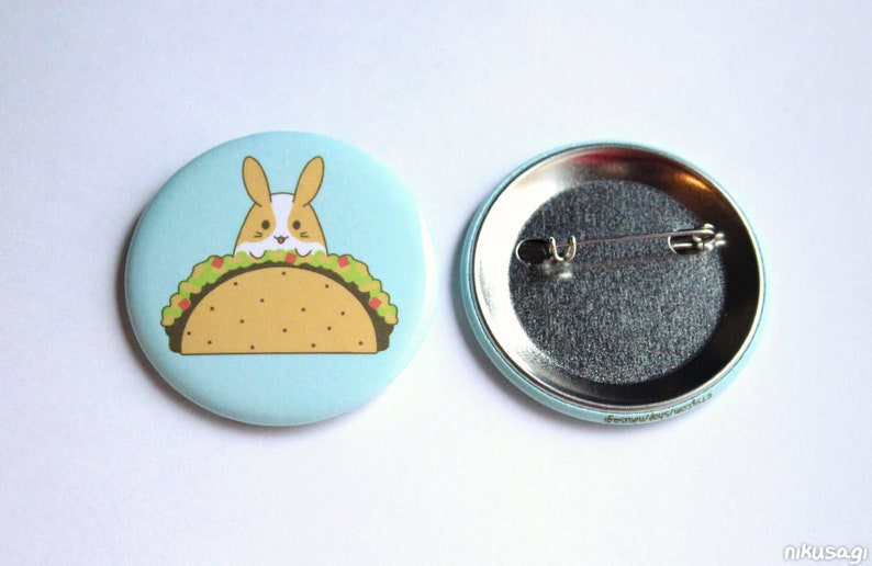 May include: A light blue button with a cartoon bunny rabbit peeking out of a taco. The back of the button is silver with a pin.