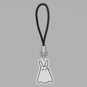 Puede incluir: Un colgante de conejito blanco con contorno negro y una cara sonriente, sujeto a un cordón negro. El colgante tiene un diseño sencillo de estilo caricatura. El cordón está conectado a un cierre metálico plateado.