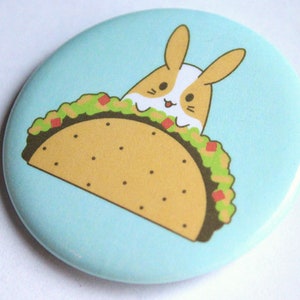 May include: A light blue button with a cartoon illustration of a brown and white rabbit peeking out of a taco. The taco is filled with green guacamole and red tomatoes.