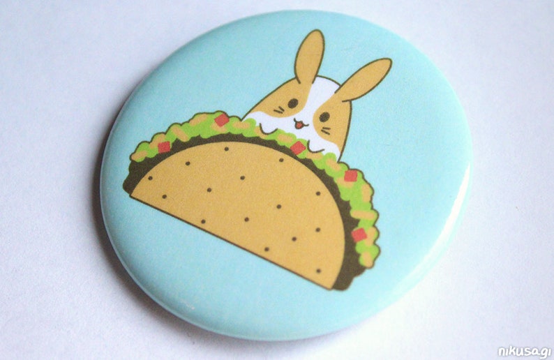 May include: A light blue button with a cartoon illustration of a brown and white rabbit peeking out of a taco. The taco is filled with green guacamole and red tomatoes.