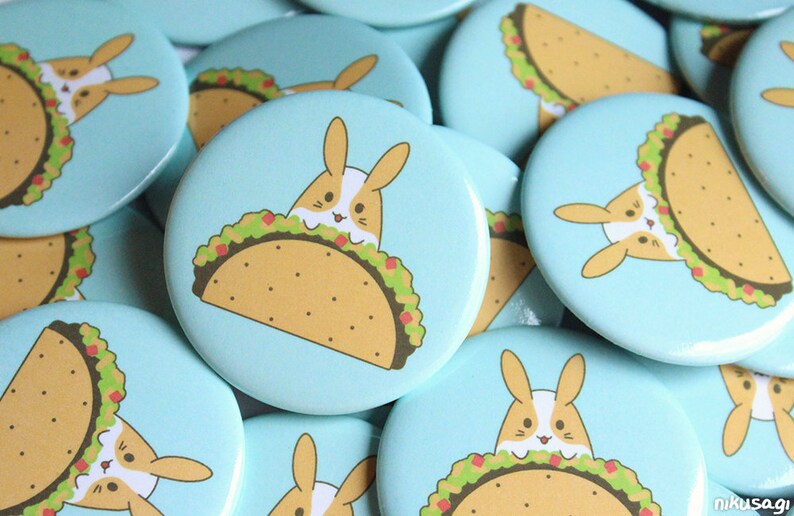 May include: A collection of blue and white round buttons with a cartoon image of a bunny rabbit peeking out of a taco. The taco is filled with lettuce, tomatoes, and cheese.