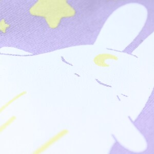 May include: A white bunny with a crescent moon on its forehead is sleeping on a lavender background with yellow stars.