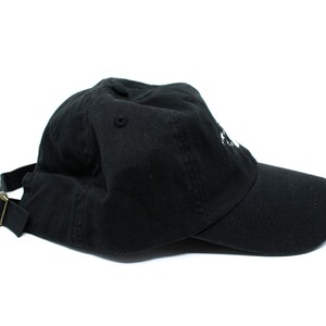 May include: A black baseball cap with a curved brim and adjustable strap. The cap has a small, white embroidered design on the front. The strap has a brass-colored metal buckle. The cap is made of a durable fabric.
