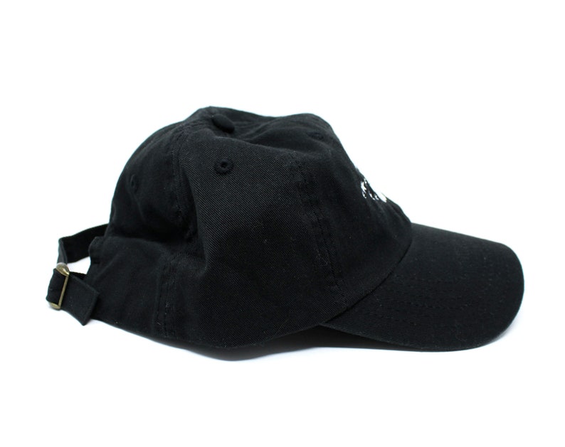 May include: A black baseball cap with a curved brim and adjustable strap. The cap has a small, white embroidered design on the front. The strap has a brass-colored metal buckle. The cap is made of a durable fabric.