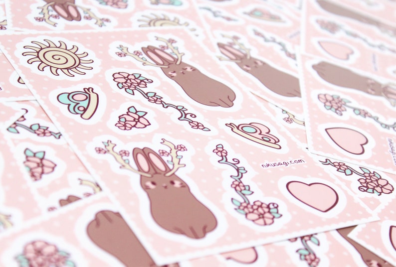 Może przedstawiać: Zestaw r&oacute;żowych i brązowych naklejek przedstawiających uroczego kr&oacute;liczka z rogami, kwiaty, serce i słońce. Naklejki znajdują się na r&oacute;żowym tle w białe kropki. Tekst "nikusagi.com" jest nadrukowany na jednej z naklejek.