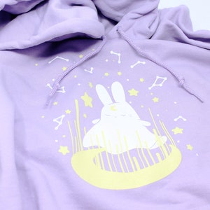May include: A lavender hoodie with a white bunny sitting on a yellow crescent moon. The bunny has a crescent moon on its forehead and is surrounded by stars.