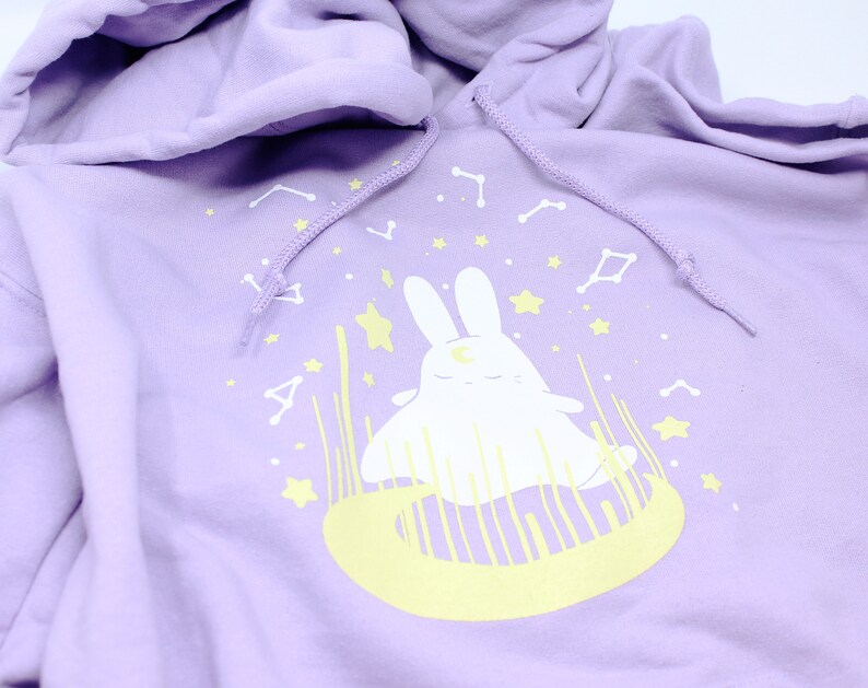 May include: A lavender hoodie with a white bunny sitting on a yellow crescent moon. The bunny has a crescent moon on its forehead and is surrounded by stars.