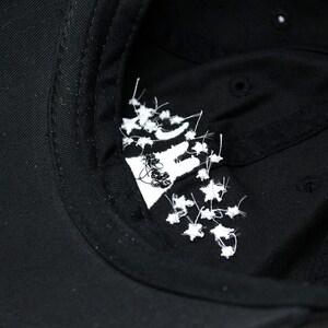 May include: Black baseball cap with a white embroidered design on the inside. The design appears to be a stylized image with white star-like accents. The cap is made of a dark fabric.