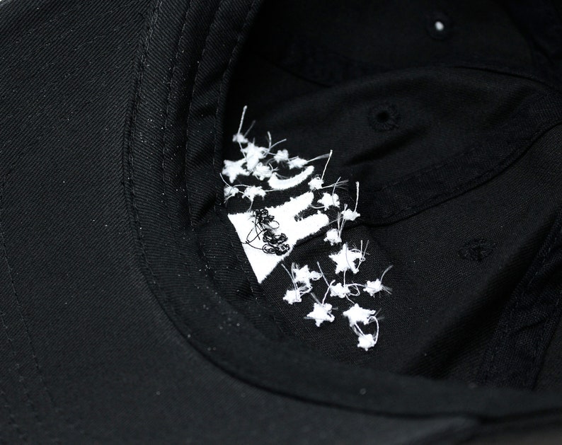 May include: Black baseball cap with a white embroidered design on the inside. The design appears to be a stylized image with white star-like accents. The cap is made of a dark fabric.