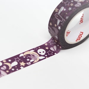 May include: A roll of decorative washi tape with a dark purple background and a repeating pattern of stars, moons, bats, skulls, and broken hearts. The tape is partially unrolled, revealing the design. The brand name is visible on the roll.
