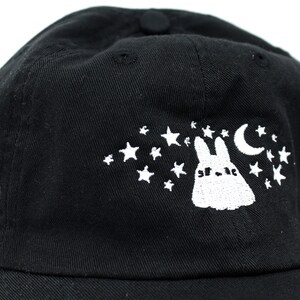 May include: Black baseball cap featuring a white embroidered design of a rabbit, stars, and a crescent moon. The cap has a curved brim and a button on top. The design is on the front of the cap.