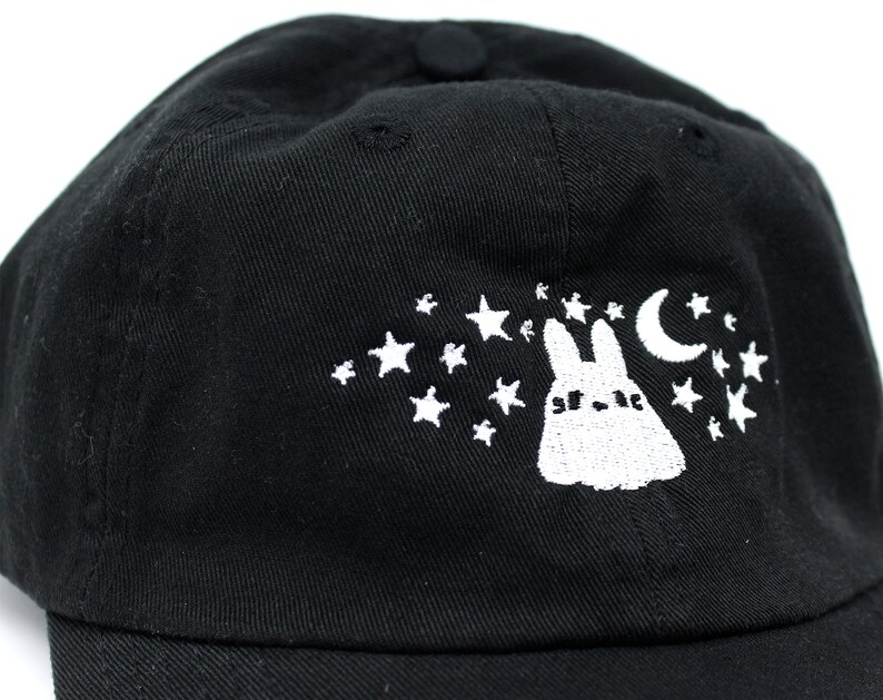 May include: Black baseball cap featuring a white embroidered design of a rabbit, stars, and a crescent moon. The cap has a curved brim and a button on top. The design is on the front of the cap.