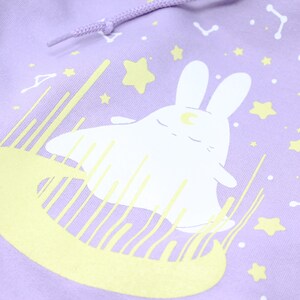 May include: A lavender hoodie with a white bunny rabbit wearing a crescent moon on its head. The bunny is surrounded by yellow stars and a yellow crescent moon.