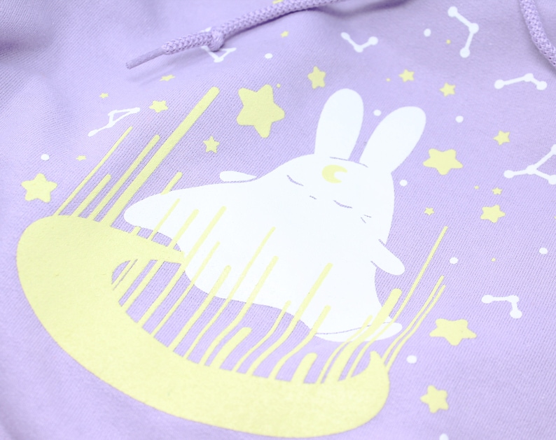 May include: A lavender hoodie with a white bunny rabbit wearing a crescent moon on its head. The bunny is surrounded by yellow stars and a yellow crescent moon.