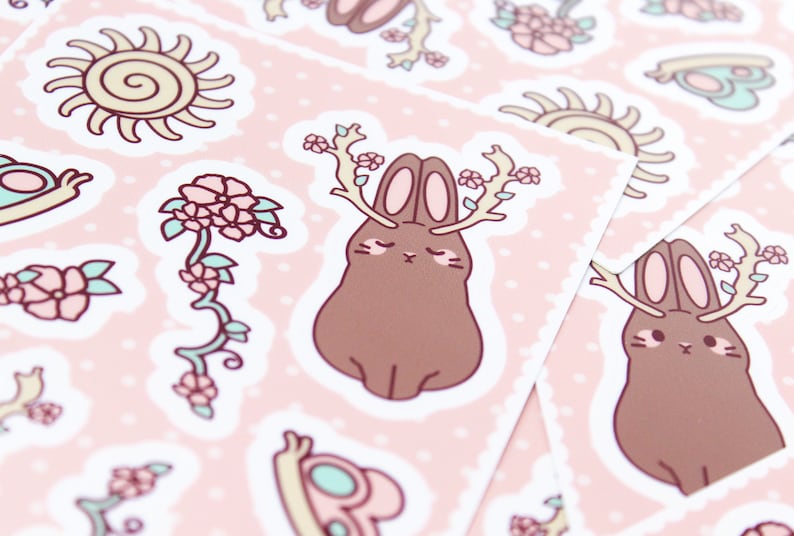 Może przedstawiać: Arkusz naklejek z brązowym kr&oacute;likiem z rogami, r&oacute;żowymi kwiatami, ż&oacute;łtym słońcem i zieloną żabą. Naklejki są na r&oacute;żowym tle z białymi kropkami.