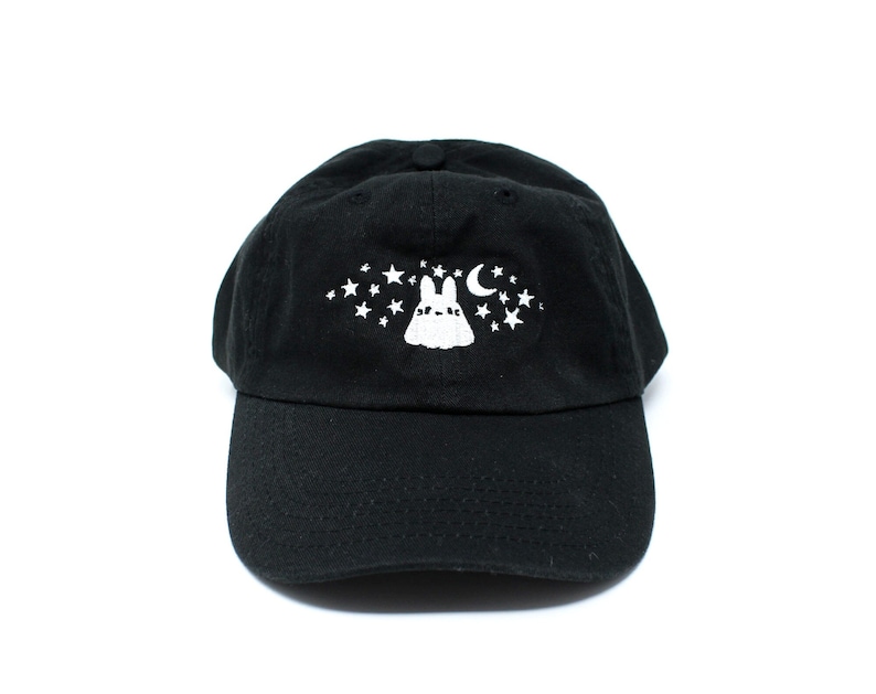 May include: Black baseball cap with a white embroidered design featuring a bunny, stars, and a crescent moon. The cap has a curved brim and an adjustable strap at the back. The hat is made of a soft, durable fabric.