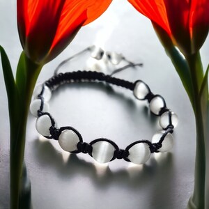 以下が含まれることがあります： 黒い編み紐に白いビーズをあしらったブレスレット。丸く磨かれた白いビーズが特徴です。背景には赤いチューリップと反射面があり、柔らかく拡散した光を作り出しています。