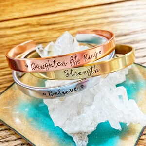 May include: Three stacked metal bracelets with inspirational messages. The top bracelet is copper and says "Daughter of the King". The middle bracelet is gold and says "Strength". The bottom bracelet is silver and says "Believe".