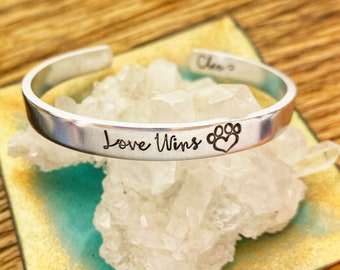 May include: Silver bracelet with the inscription "Love Wins" and a paw print with a heart in the center.