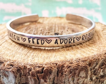 May include: Silver bracelet with the words "Latch Feed Adore Repeat" engraved on it with a heart symbol between each word.