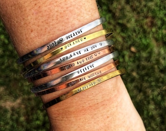 May include: A stack of seven silver, gold, and copper bracelets with inspirational messages engraved on them. The messages include "Just Breathe", "Dreamer", "God Gave Me You", "Wild and Free", "Believe", "You Are Worthy", and "Redeemed".