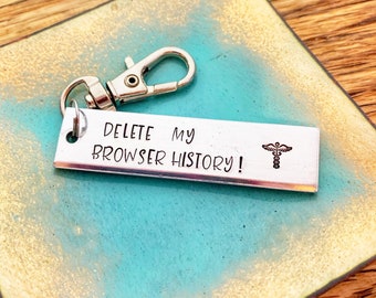 May include: Silver keychain with a silver rectangle charm that says "DELETE MY BROWSER HISTORY!" with a medical symbol on the right side. The keychain is on a turquoise and gold background.