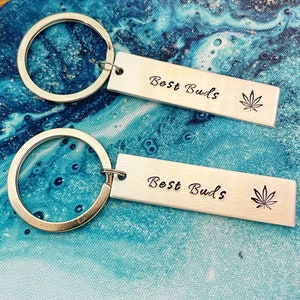 Puede incluir: Dos llaveros plateados con un anillo plateado y una etiqueta rectangular. Cada etiqueta tiene el texto "Best Buds" grabado con una hoja de marihuana estilizada.