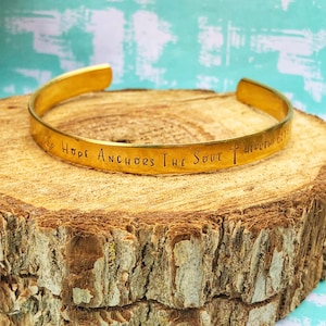 Puede incluir: Pulsera de metal dorado con la inscripción "Hope Anchors The Soul Hebrews 6:19" sobre una superficie de madera.