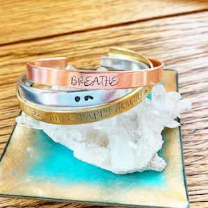 Puede incluir: Tres pulseras de metal apiladas con mensajes inspiradores: "Respira", "Piensa en pensamientos felices" y un punto y coma. Las pulseras están sobre una roca de cristal blanco.