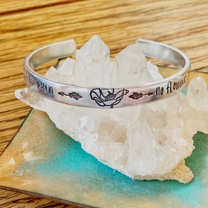 May include: A silver cuff bracelet with the words "no pain no flowers" engraved on the outside. The bracelet features a floral design and arrows. The bracelet is displayed on a bed of clear crystals.