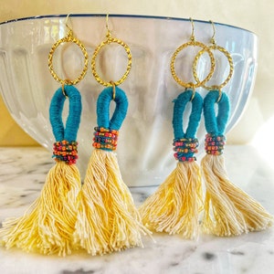 May include: A pair of gold hoop earrings with turquoise and multi-colored beaded tassels. The tassels are made of a soft, fluffy material.