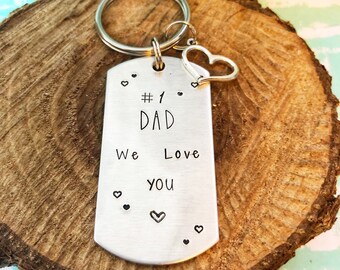 Puede incluir: Llavero de plata con un dije de corazón y una etiqueta que dice "#1 DAD We Love You" con pequeños corazones alrededor del texto.