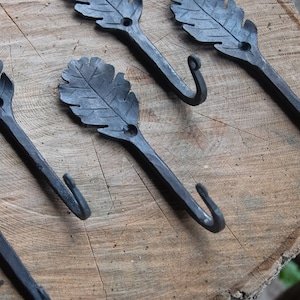 Puede incluir: Cinco ganchos de metal negro con forma de hojas de roble. Los ganchos están dispuestos en una fila sobre una superficie de madera.