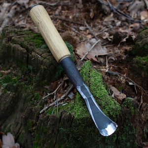 Może przedstawiać: Narzędzie do rzeźbienia z drewnianym uchwytem i ostrzem z czarnego metalu, leżące na porośniętym mchem pniu drzewa w lesie.