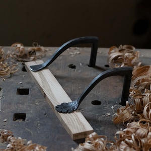 May include: Two black metal handles with leaf-shaped details are attached to a wooden board. The board is surrounded by wood shavings on a wooden workbench.