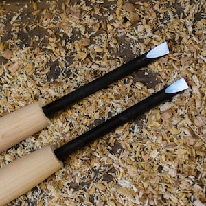 Può includere: Due scalpelli con manico in legno, lame in metallo nero e punte bianche. Gli scalpelli sono appoggiati su una superficie ricoperta di trucioli di legno.