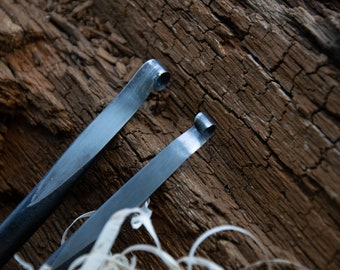 May include: Two metal tools with curved ends, lying on a piece of weathered wood. The tools are made of a dark metal and have a shiny finish.
