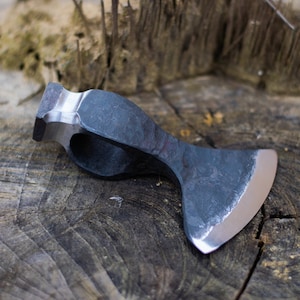 Op de afbeelding: Een handgesmede bijlkop met een donkere, getextureerde afwerking en een gepolijst zilveren blad. De bijlkop rust op een verweerd houten oppervlak, wat het vakmanschap en de rustieke uitstraling laat zien. Een gereedschap voor houtbewerking en buitenactiviteiten.