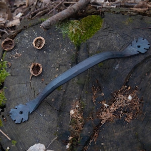 May include: A black metal door handle with a decorative, leaf-shaped design on each end. The handle is lying on a rough, wooden surface.