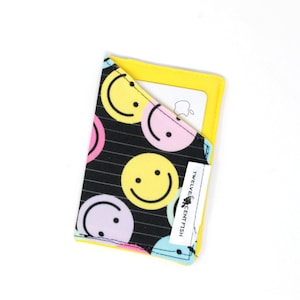 May include: A fabric card holder with a black background and colorful smiley faces. The interior is yellow, and a white label with text is visible. A card is partially visible in the pocket.